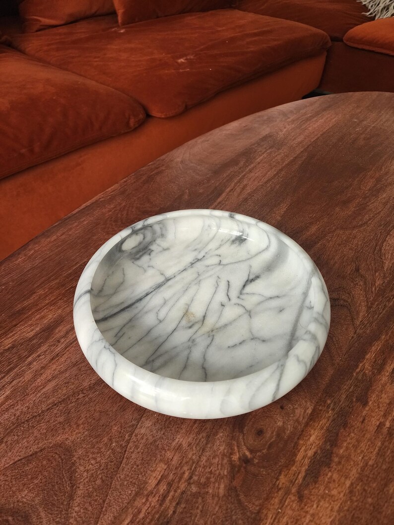 May include: A round, shallow bowl crafted from white marble with prominent grey veining throughout. The bowl features a smooth, polished surface and a thick, rounded rim, showcasing the natural stone patterns and variations in the marble material.