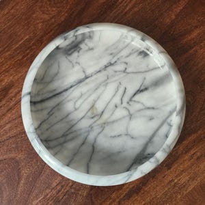 May include: A circular white marble tray featuring prominent grey veining throughout the surface. The tray has a smooth, polished finish and a raised, rounded rim that creates a shallow basin, showcasing the natural stone pattern and texture.