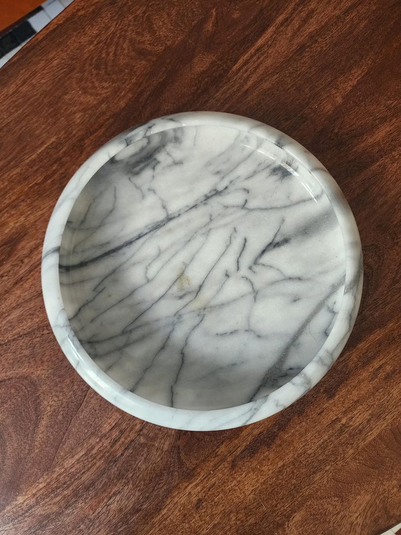 May include: A circular white marble tray featuring prominent grey veining throughout the surface. The tray has a smooth, polished finish and a raised, rounded rim that creates a shallow basin, showcasing the natural stone pattern and texture.