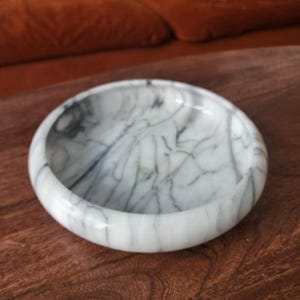 May include: A circular white marble bowl featuring prominent dark gray veining throughout its surface. The bowl has a smooth, polished finish with a thick, rounded rim and a shallow interior basin, showcasing the natural stone pattern.