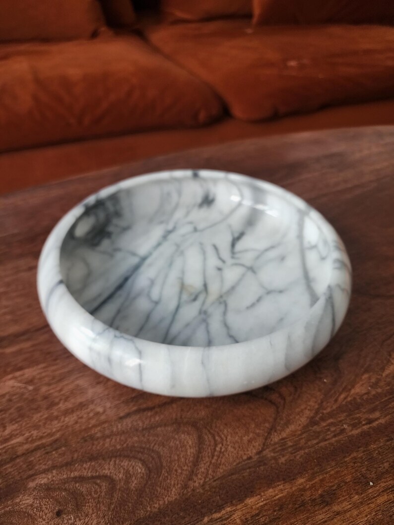 May include: A circular white marble bowl featuring prominent dark gray veining throughout its surface. The bowl has a smooth, polished finish with a thick, rounded rim and a shallow interior basin, showcasing the natural stone pattern.