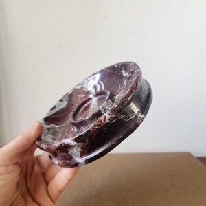 May include: A circular dish crafted from polished dark burgundy marble with prominent white veining. The object features a smooth, concave surface with a raised central circular platform, showcasing a natural stone pattern throughout the entire piece.