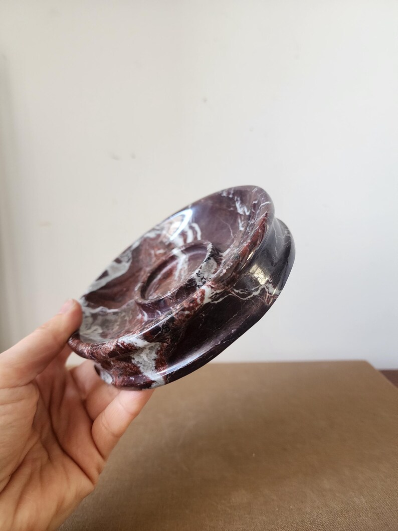 May include: A circular dish crafted from polished dark burgundy marble with prominent white veining. The object features a smooth, concave surface with a raised central circular platform, showcasing a natural stone pattern throughout the entire piece.