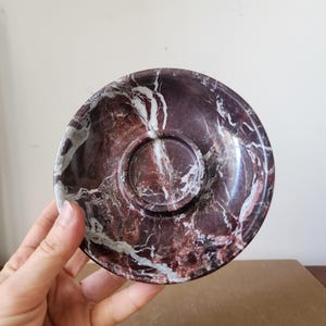 May include: A circular saucer crafted from polished dark maroon marble with prominent white veining throughout. The saucer features a raised central ring designed to hold a cup securely in place, showcasing a smooth, glossy stone finish.