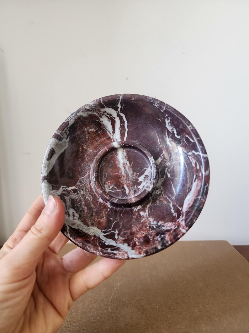 May include: A circular saucer crafted from polished dark maroon marble with prominent white veining throughout. The saucer features a raised central ring designed to hold a cup securely in place, showcasing a smooth, glossy stone finish.