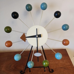 May include: A sunburst-style clock featuring a central white circular face with black hands and a brown triangular pointer. Twelve thin metal spokes extend outwards, each tipped with a sphere in various colours including blue, green, black, and orange.