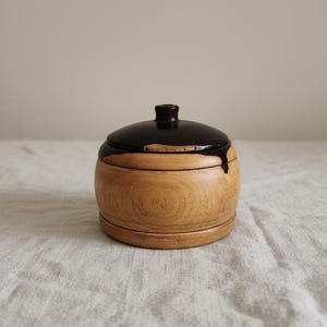 Puede incluir: Un pequeño recipiente de madera redondo con una tapa marrón oscuro. La tapa tiene un pequeño pomo en la parte superior y una gota del material marrón oscuro en el lateral. El recipiente es de madera clara con vetas visibles.