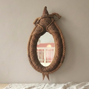 May include: Oval-shaped mirror with a woven rope frame. The frame is a light brown color and has a decorative top element. The mirror hangs on a neutral-colored wall. The mirror's shape is reminiscent of a saddle.