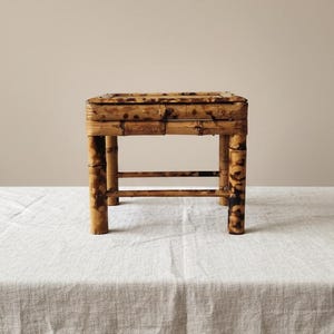 May include: A small, square bamboo stool with a natural finish. The stool has four legs and a slatted top. The bamboo is a warm brown colour with darker accents. The stool is approximately 30 cm tall.