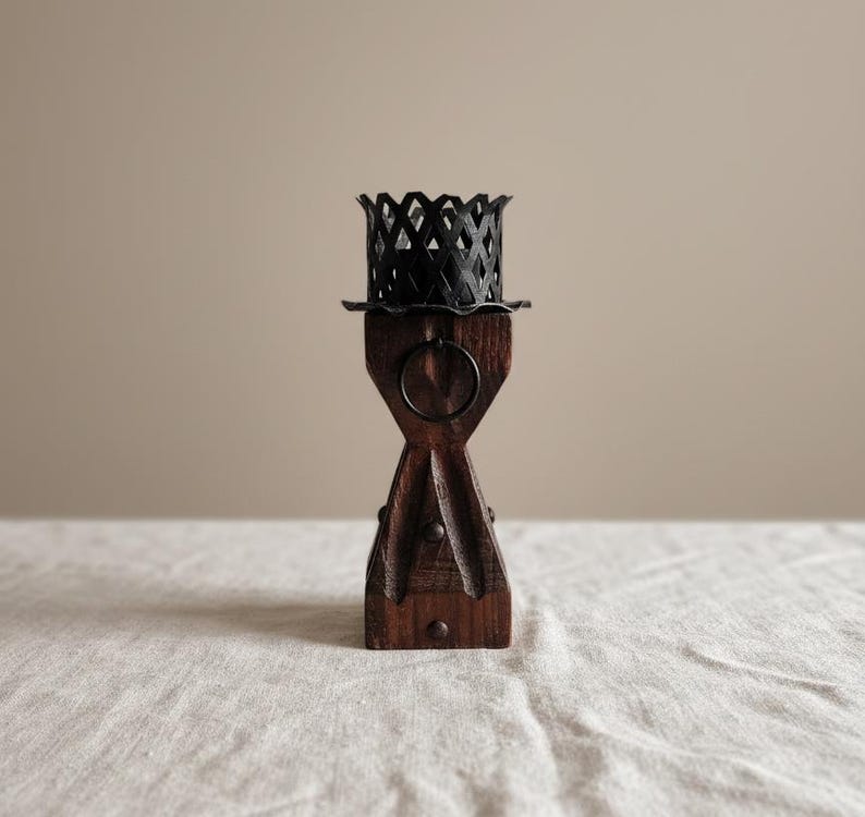 May include: A dark brown wooden candle holder featuring a tapered, hourglass-shaped base with vertical grooves and a small metal ring detail. The top is crowned with a black metal lattice cup designed to hold a candle securely in place.