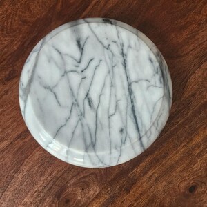 May include: A circular white marble tray featuring prominent grey veining throughout its surface. The object has a smooth, polished finish and a slightly raised edge, presenting a consistent pattern of natural stone markings across the entire top.