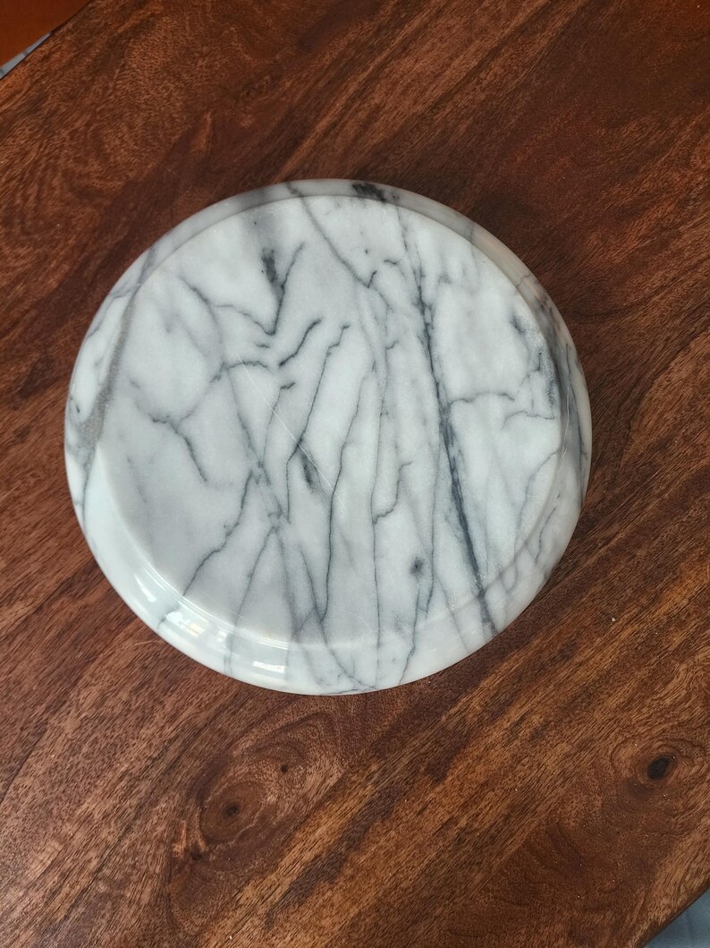 May include: A circular white marble tray featuring prominent grey veining throughout its surface. The object has a smooth, polished finish and a slightly raised edge, presenting a consistent pattern of natural stone markings across the entire top.