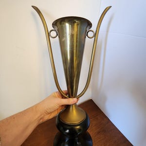 Pode incluir: Uma ta&ccedil;a de trof&eacute;u de lat&atilde;o alta com uma borda alargada, duas al&ccedil;as finas e curvas, e um acabamento polido. O corpo de metal est&aacute; montado sobre uma base preta s&oacute;lida e arredondada. Texto gravado &eacute; vis&iacute;vel na frente da ta&ccedil;a.