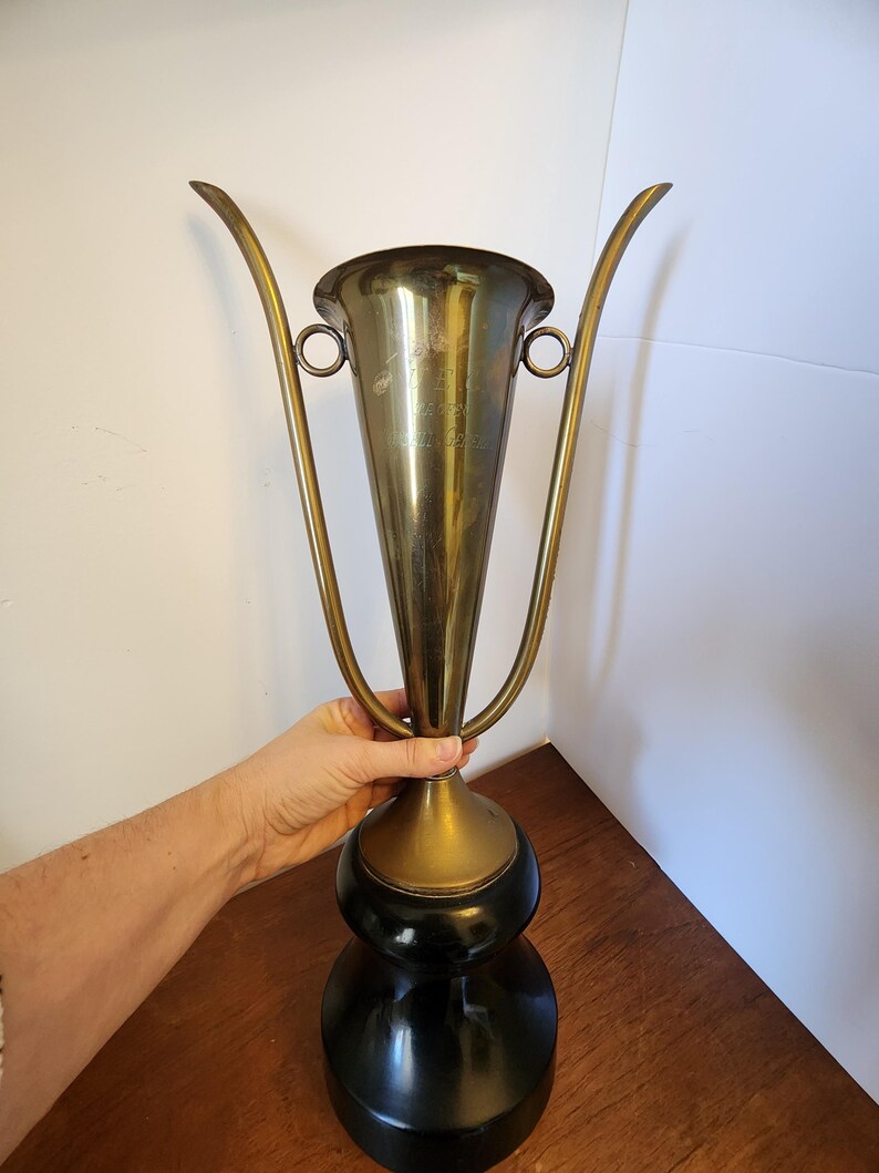 Pode incluir: Uma ta&ccedil;a de trof&eacute;u de lat&atilde;o alta com uma borda alargada, duas al&ccedil;as finas e curvas, e um acabamento polido. O corpo de metal est&aacute; montado sobre uma base preta s&oacute;lida e arredondada. Texto gravado &eacute; vis&iacute;vel na frente da ta&ccedil;a.