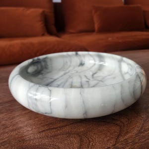 May include: A circular white marble bowl featuring prominent grey veining throughout its polished surface. The bowl has a thick, rounded rim and a shallow interior basin, showcasing the natural stone pattern with a smooth, glossy finish.