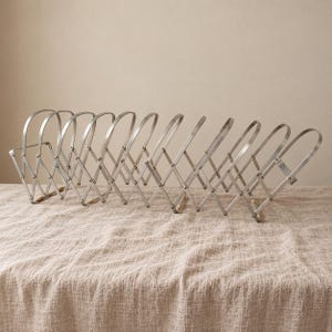 Puede incluir: Un estante extensible de metal plateado que presenta una serie de divisores arqueados conectados por un mecanismo de tijera. La estructura está construida con tiras de metal delgadas y planas e incluye pequeños detalles de madera.