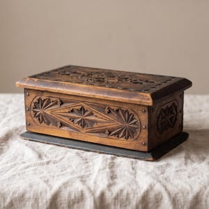 May include: A rectangular wooden box with a dark brown finish, featuring intricate geometric star-shaped carvings on the lid and front panel. The box is constructed with visible metal nail heads along the edges and sits on a dark base.