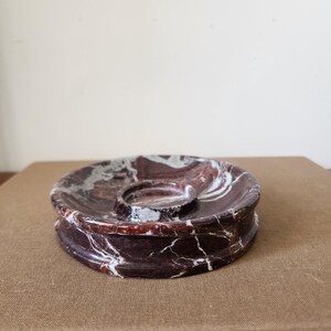 May include: A circular dark red and white marble tray featuring a central raised circular indentation. The stone surface displays natural veining patterns throughout the deep burgundy and white color palette, creating a polished and smooth finish.