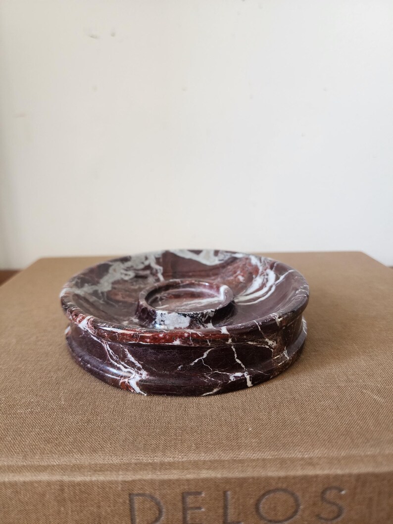 May include: A circular dark red and white marble tray featuring a central raised circular indentation. The stone surface displays natural veining patterns throughout the deep burgundy and white color palette, creating a polished and smooth finish.