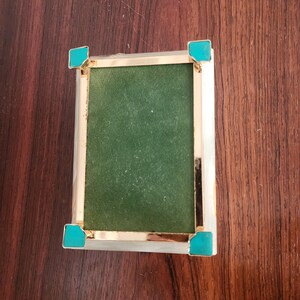 Puede incluir: Una caja rectangular con un marco de metal dorado pulido y cuatro acentos de esquina en color turquesa brillante. El panel central est&aacute; cubierto con una tela verde oscuro texturizada y los bordes exteriores tienen un acabado blanco liso.