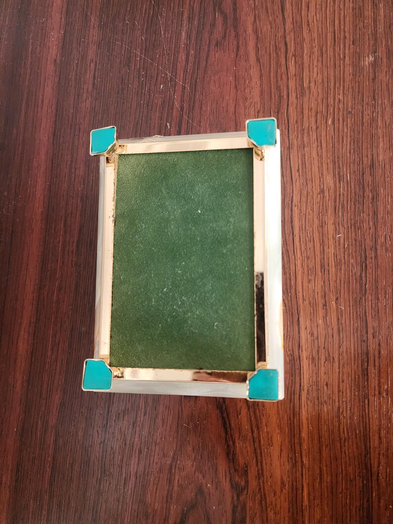 Puede incluir: Una caja rectangular con un marco de metal dorado pulido y cuatro acentos de esquina en color turquesa brillante. El panel central est&aacute; cubierto con una tela verde oscuro texturizada y los bordes exteriores tienen un acabado blanco liso.