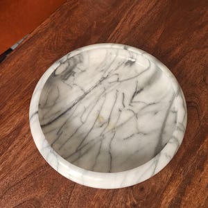 May include: A circular white marble bowl featuring prominent grey veining throughout the stone surface. The bowl has a thick, rounded rim and a smooth, polished finish, showcasing the natural patterns and variations inherent in the marble material.