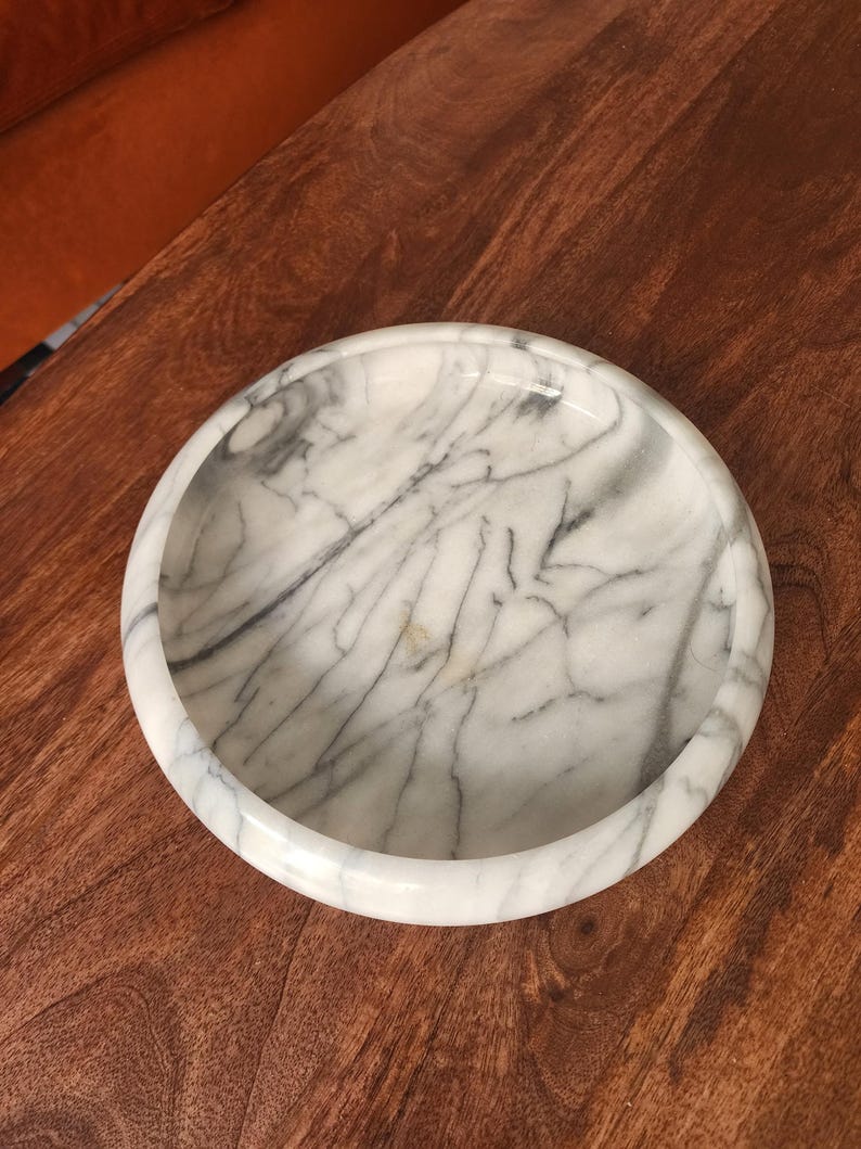 May include: A circular white marble bowl featuring prominent grey veining throughout the stone surface. The bowl has a thick, rounded rim and a smooth, polished finish, showcasing the natural patterns and variations inherent in the marble material.