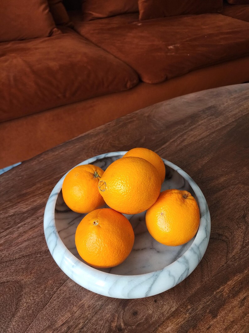 May include: A round, white marble bowl with grey veining containing five bright orange citrus fruits. The bowl has a smooth, polished finish and a thick, rounded rim, providing a stark contrast to the vibrant, textured skins of the oranges.