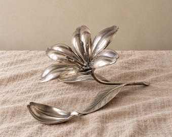 May include: A silver-toned metal sculpture shaped like a blooming lily flower with a long stem and a single detached petal resting nearby. The metallic surface has a polished finish with visible veins on the leaf and petal structures.