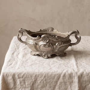 May include: A silver-toned metal gravy boat or sauce tureen featuring ornate, raised scrollwork and floral relief detailing. The vessel has two curved handles on opposite sides and rests on a decorative, scalloped base with four small feet.