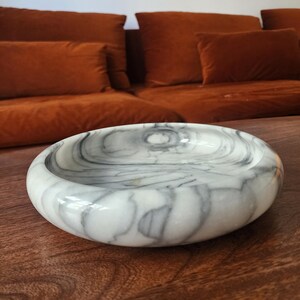 May include: A circular bowl crafted from white stone with prominent grey veining throughout its surface. The bowl features a smooth, polished finish and a thick, rounded rim, creating a shallow basin for holding small items or decorative display.