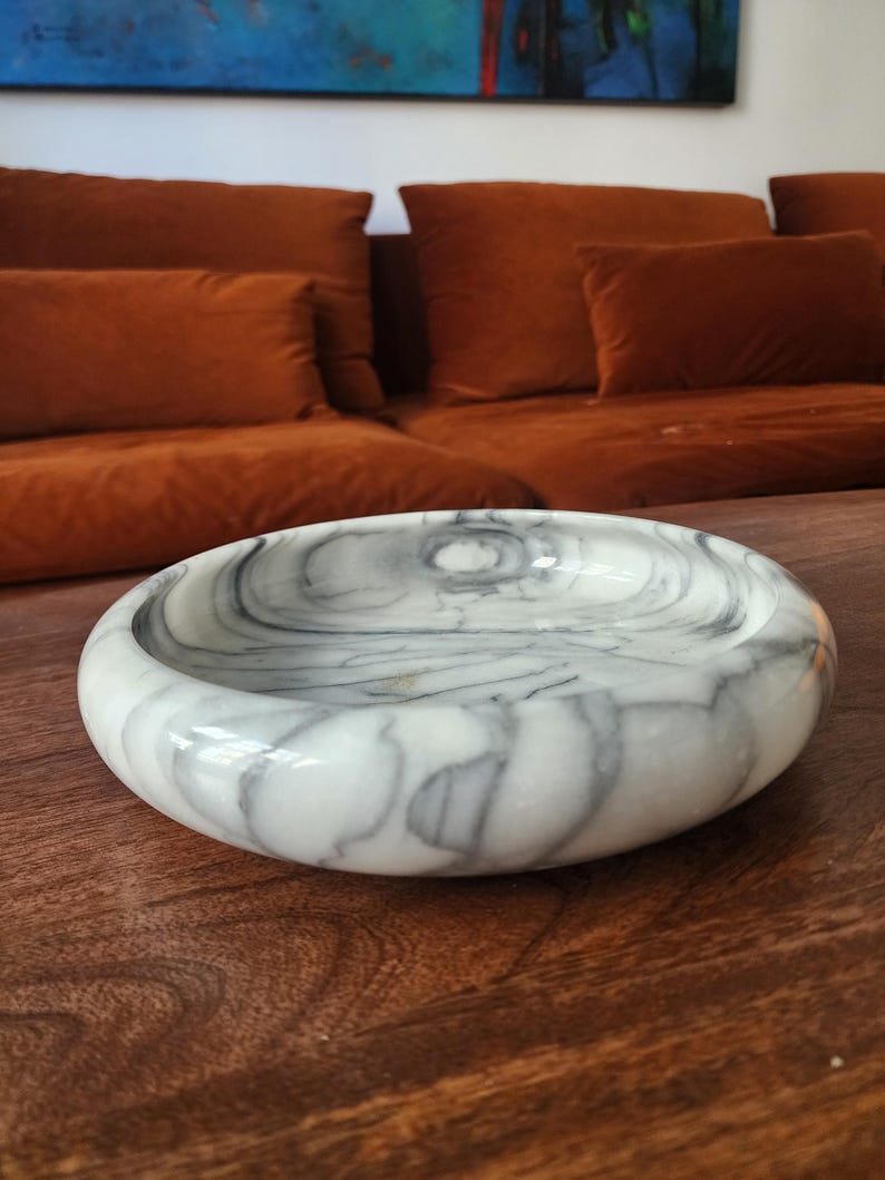 May include: A circular bowl crafted from white stone with prominent grey veining throughout its surface. The bowl features a smooth, polished finish and a thick, rounded rim, creating a shallow basin for holding small items or decorative display.