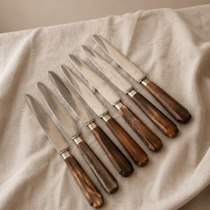 Könnte beinhalten: Ein Set aus sechs Tafelmessern, die in einer diagonalen Reihe angeordnet sind. Jedes Messer verfügt über eine polierte, silberfarbene Metallklinge und einen facettierten, dunkelbraunen Griff mit einer holzähnlichen Maserung.