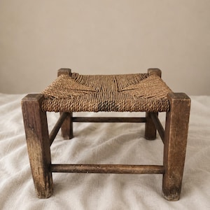 May include: A small, low stool featuring four weathered, dark brown wooden legs connected by horizontal support bars. The square seat is constructed from tightly woven, natural tan fibre cord, creating a textured, crisscross pattern across the top.