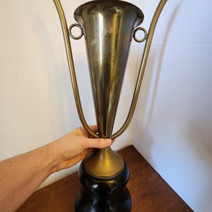 Pode incluir: Um trof&eacute;u alto de metal dourado com uma borda alargada e duas al&ccedil;as longas e curvas que se estendem para cima. A ta&ccedil;a apresenta texto gravado na frente e est&aacute; montada sobre uma base preta s&oacute;lida, lisa e arredondada com acabamento polido.
