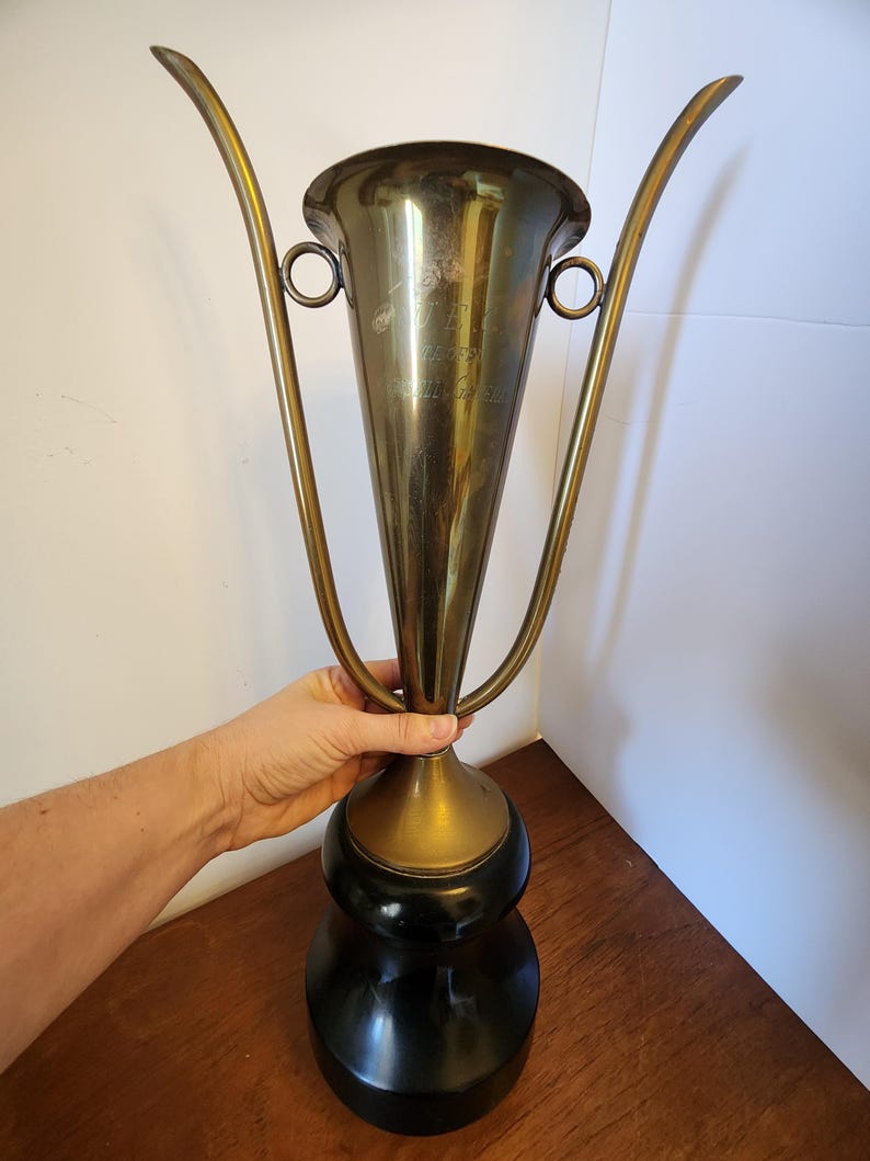 Pode incluir: Um trof&eacute;u alto de metal dourado com uma borda alargada e duas al&ccedil;as longas e curvas que se estendem para cima. A ta&ccedil;a apresenta texto gravado na frente e est&aacute; montada sobre uma base preta s&oacute;lida, lisa e arredondada com acabamento polido.