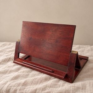 Può includere: Un leggio in legno marrone rossastro scuro con un pannello posteriore rettangolare, una sporgenza orizzontale per appoggiare i libri e due piccole cerniere in metallo color ottone che collegano la struttura di supporto alla base.