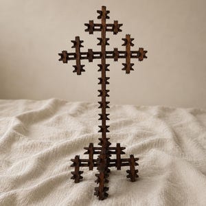 Puede incluir: Una cruz de madera de color marrón oscuro con un diseño de tres barras, que presenta una serie de pequeños elementos decorativos en forma de estrella dentada a lo largo de los bordes del poste vertical y las tres barras transversales, sobre una base de cuatro patas.