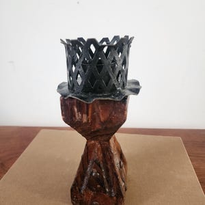 May include: A candle holder featuring a dark brown, textured wooden base with a tapered, geometric shape and visible metal studs. At the top, a black metal cup with a lattice-patterned rim and a scalloped edge sits securely on the wooden stand.
