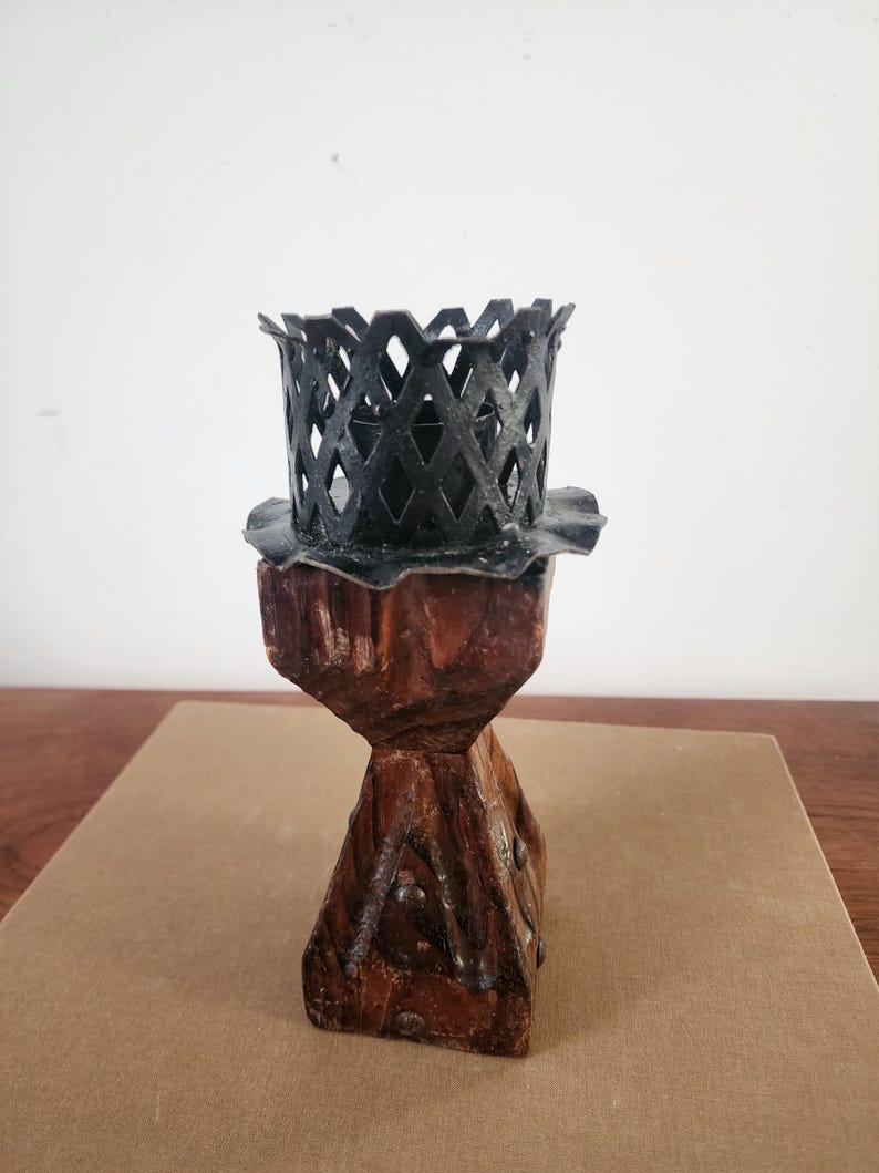 May include: A candle holder featuring a dark brown, textured wooden base with a tapered, geometric shape and visible metal studs. At the top, a black metal cup with a lattice-patterned rim and a scalloped edge sits securely on the wooden stand.