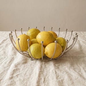Pode incluir: Uma fruteira de metal prateado cheia de vários limões amarelos. A tigela tem um design único com suportes curvos e pequenos detalhes de bolas. Os limões são de vários tamanhos e tons de amarelo.