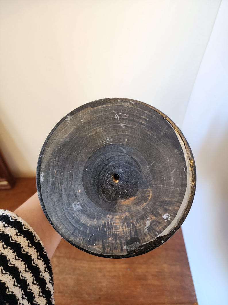 Pode incluir: Uma base circular de madeira com acabamento escuro e carbonizado, exibindo an&eacute;is de crescimento conc&ecirc;ntricos vis&iacute;veis e um pequeno orif&iacute;cio central. A borda do disco apresenta um fino acabamento met&aacute;lico dourado e desgastado ao redor.