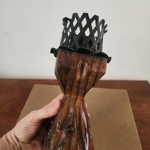 May include: A candle holder featuring a dark brown, textured wooden base with a tapered, hourglass-like shape and visible metal studs. The top is fitted with a black metal cage-like crown with a diamond-patterned lattice design.