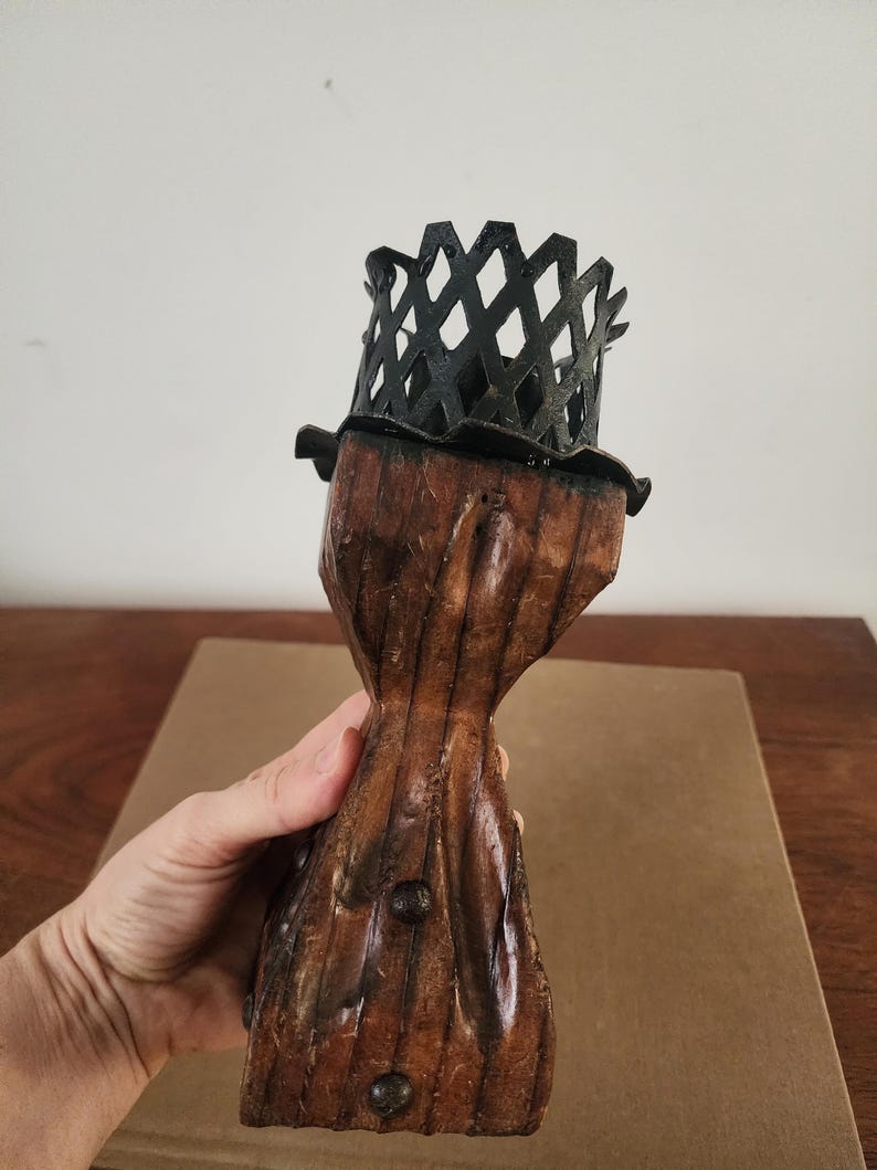 May include: A candle holder featuring a dark brown, textured wooden base with a tapered, hourglass-like shape and visible metal studs. The top is fitted with a black metal cage-like crown with a diamond-patterned lattice design.