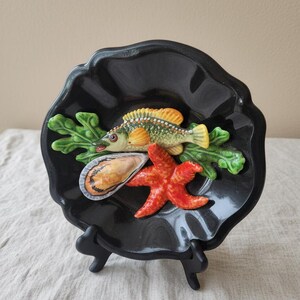 Könnte beinhalten: Ein schwarzer dekorativer Keramikteller mit einem Fisch-, Seestern- und Muscheldesign. Der Fisch ist grün, gelb und orange, der Seestern ist rot und die Muschel ist beige. Der Teller steht auf einem schwarzen Ständer.