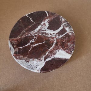 May include: A circular coaster featuring a deep burgundy and dark red marble surface with prominent, irregular white veining throughout. The stone has a polished finish, showcasing the natural color variations and intricate patterns of the marble.