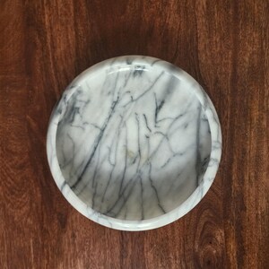 May include: A circular white marble bowl featuring prominent grey veining throughout the stone surface. The bowl has a smooth, polished finish with a thick, rounded rim and a deep, concave interior basin, viewed from directly above.