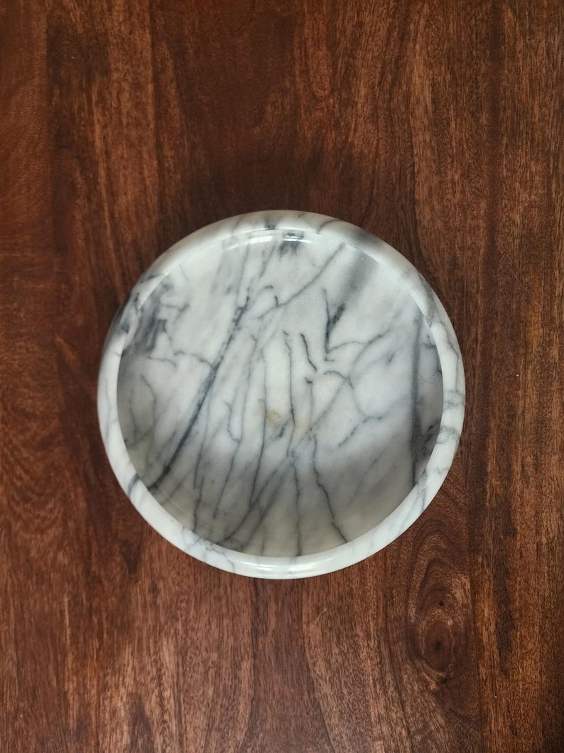 May include: A circular white marble bowl featuring prominent grey veining throughout the stone surface. The bowl has a smooth, polished finish with a thick, rounded rim and a deep, concave interior basin, viewed from directly above.