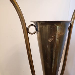 Pode incluir: Uma ta&ccedil;a de trof&eacute;u c&oacute;nica de lat&atilde;o com duas pegas curvas e alongadas fixadas nas laterais com an&eacute;is circulares. A superf&iacute;cie do lat&atilde;o apresenta sinais de oxida&ccedil;&atilde;o e desgaste, com um texto gravado t&eacute;nue vis&iacute;vel na parte frontal.