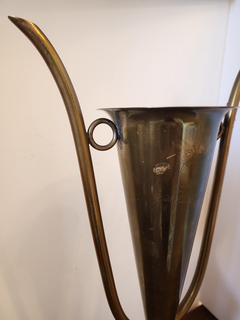 Pode incluir: Uma ta&ccedil;a de trof&eacute;u c&oacute;nica de lat&atilde;o com duas pegas curvas e alongadas fixadas nas laterais com an&eacute;is circulares. A superf&iacute;cie do lat&atilde;o apresenta sinais de oxida&ccedil;&atilde;o e desgaste, com um texto gravado t&eacute;nue vis&iacute;vel na parte frontal.